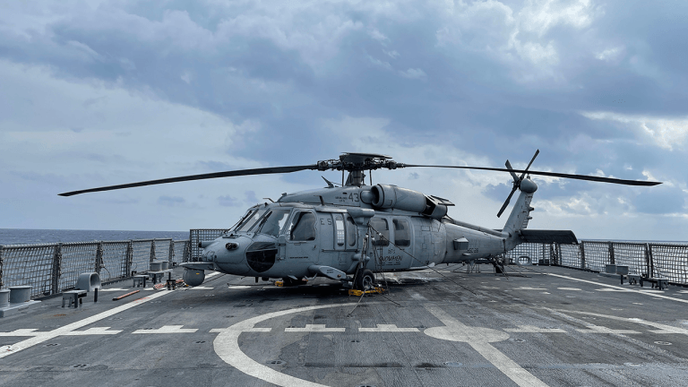 A military helicopter on a naval flight deck under a cloudy sky at sea.